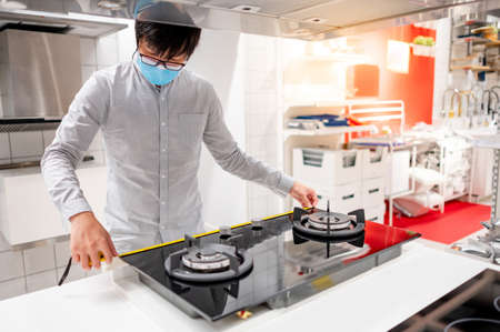 Asian man interior designer wearing face mask using tape measure on gas stove on counter in kitchen showroom. Shopping appliance for domestic kitchen. Home improvement conceptの写真素材