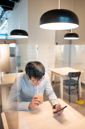 Asian man drinking coffee and using smartphone while sitting in table shield partition for...