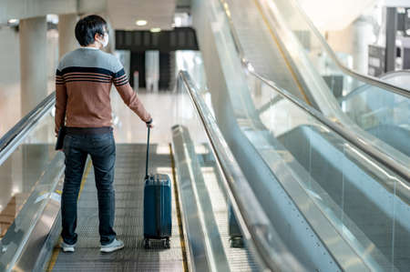 Travel insurance concept. Asian man tourist carrying suitcase luggage and digital tablet on travelator or moving walkway in airport terminal.の写真素材
