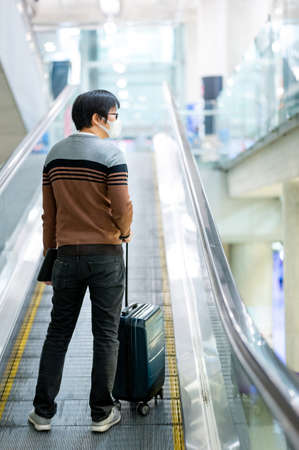 Travel insurance concept. Asian man tourist wearing face mask carrying suitcase luggage and digital tablet on travelator or moving walkway in airport terminal.の写真素材