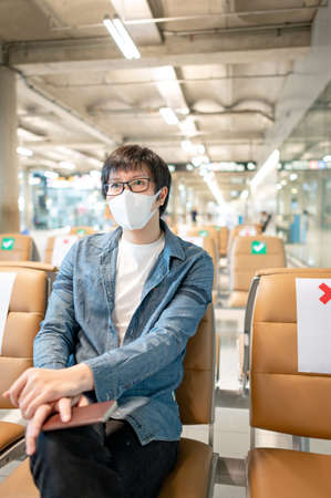 Asian man tourist wearing protective face mask holding passport sitting in airport terminal. Coronavirus (COVID-19) pandemic prevention when travel abroad. Health awareness and social distancingの写真素材