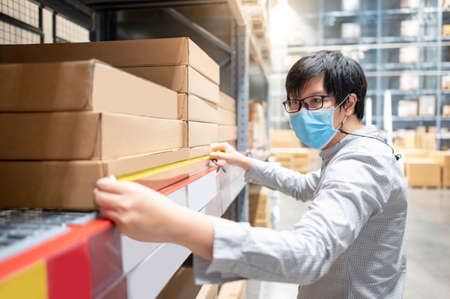 Asian man worker wearing face mask using tape measure for measuring dimension of product in cardboard box. Stock quality control in warehouse.の写真素材