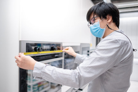 Asian man interior designer wearing face mask using tape measure on oven in kitchen showroom at...