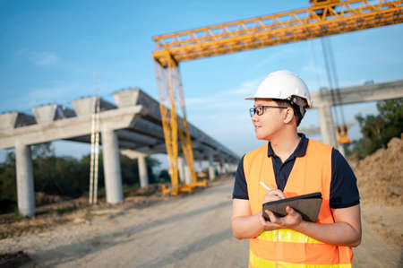 Smart Asian worker man or male civil engineer with protective safety helmet and reflective vest using digital tablet for project planning and checking architectural drawing at construction site.の写真素材