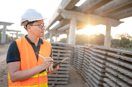 Smart Asian worker man or male civil engineer with protective safety helmet and reflective vest using digital tablet for project planning and checking material at construction site.の写真素材