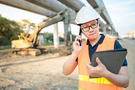 Asian worker man or male civil engineer with protective safety helmet and reflective vest using smartphone and digital tablet for project planning and checking schedule at construction site.の写真素材