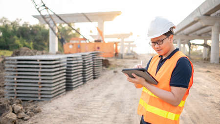 Smart Asian worker man or male civil engineer with protective safety helmet and reflective vest using digital tablet for project planning and checking architectural drawing at construction site.の写真素材