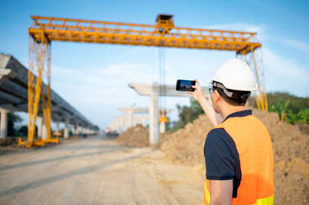 Smart Asian worker man or male civil engineer with protective safety helmet and reflective vest using using smartphone for taking photo at construction site.の写真素材