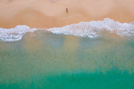 Aerial view of turquoise ocean wave reaching the coastline with tourist couple waking on the beach....