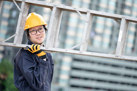 Asian maintenance worker man with protective suit and safety helmet carrying aluminium step ladder at construction site. Civil engineering, Architecture builder and building service conceptsの写真素材