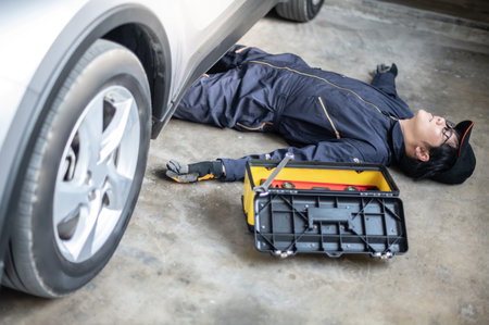 Asian auto mechanic feeling tired while working under car in auto service garage. Mechanical maintenance engineer working in automotive industry. Automobile servicing and repair conceptの写真素材
