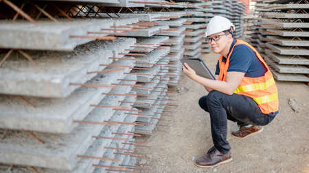 Smart Asian worker man or male civil engineer with protective safety helmet and reflective vest using digital tablet for project planning and checking material at construction site.の写真素材
