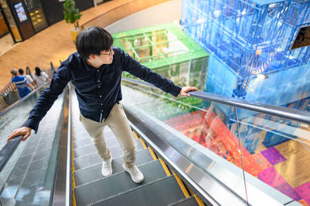 Asian man in casual clothing walking up on escalator in shopping mall or department store. Urban lifestyle in the city.の写真素材