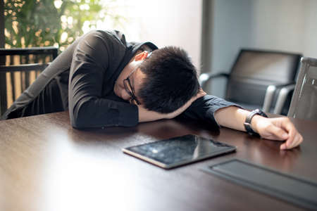 Overworked Asian businessman sleeping in office meeting room with digital tablet on conference table. Exhaustion and health problems from overload working conceptの写真素材