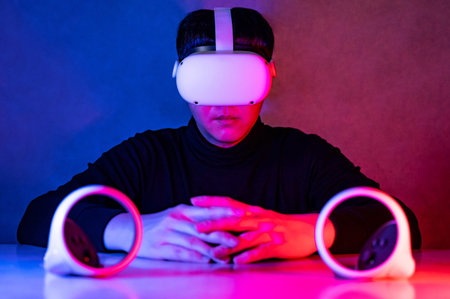 Gamer wearing virtual reality goggles sitting with controllers playing video games in neon light...
