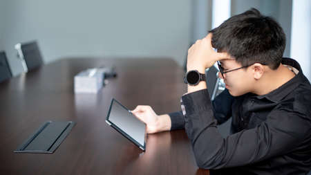 Overworked Asian Businessman feeling stressed, tired and headache while reading on digital tablet in meeting room. Depression from business problem. Mental health illness or office syndrome conceptの写真素材