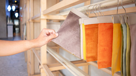 Stylist hand choosing brown fabric piece from fabric swatch material. Upholstery sample collection hanging on showcase rack. Textile industry for home interior design and furniture finishing decorの写真素材