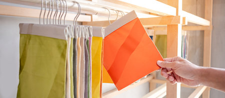 Stylist hand choosing orange fabric piece from fabric swatch material. Upholstery sample collection hanging on showcase rack. Textile industry for home interior design and furniture finishing decorの写真素材