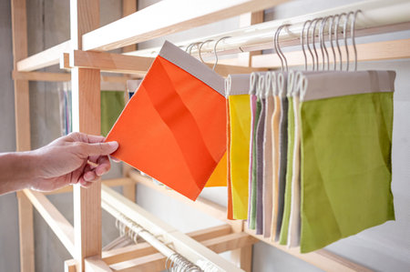 Stylist hand choosing orange fabric piece from fabric swatch material. Upholstery sample collection hanging on showcase rack. Textile industry for home interior design and furniture finishing decorの写真素材