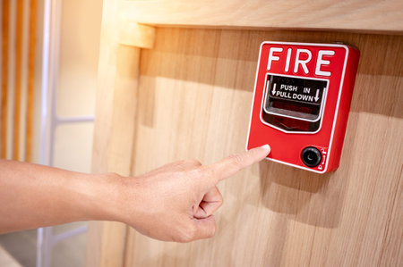 Hand pointing at red fire alarm switch mounted on wooden wall in the office interior. Fire warning system equipment for emergency and evacuation in public buildingの写真素材