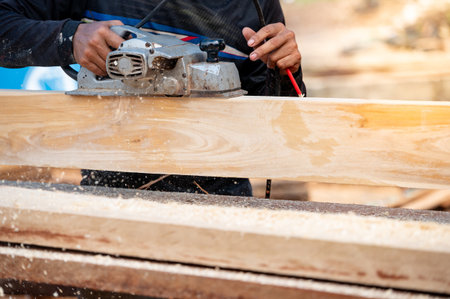 Male carpenter hand using an electric planer on wooden plank at construction site. Woodworker planing timber in his workshop with sawdust spreading around. Lumber industry and carpentry work toolの写真素材
