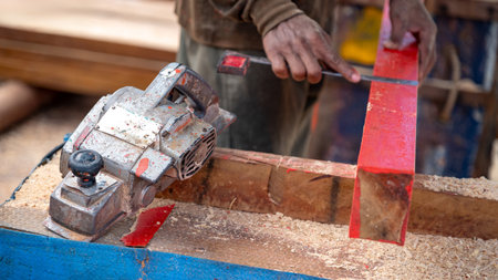 Rustic electric planer on workbench with carpenter hand squaring timber behind. Woodworker planing wooden plank at construction site. Carpentry work tool and lumber industry conceptの写真素材