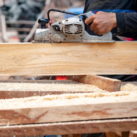 Male carpenter hand using an electric planer on wooden plank in workshop. Woodworker planing timber at construction site with sawdust spreading around. Work tool for carpentry. Lumber industry conceptの写真素材