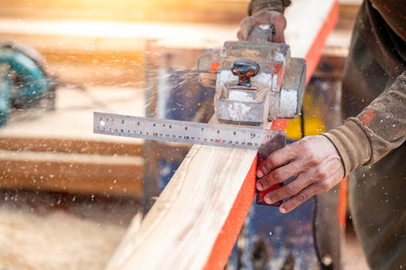 Male carpenter hand using angle ruler for wood squaring and planing on wooden plank by electric planer on workbench in workshop. Wood measuring and shaving process for lumber industryの写真素材