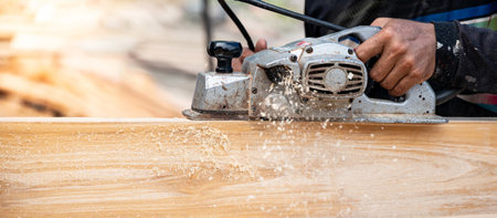 Male carpenter hand using an electric planer on wooden plank at construction site. Woodworker planing timber in his workshop with sawdust spreading around. Lumber industry and carpentry work toolの写真素材