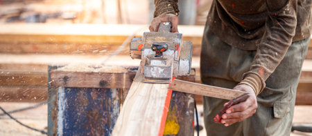 Male carpenter hand using angle ruler for wood squaring and planing on wooden plank by electric planer on workbench in workshop. Wood measuring and shaving process for lumber industryの写真素材