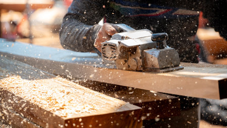 Male carpenter hand using an electric planer on wooden plank at construction site. Woodworker planing timber in his workshop with sawdust spreading around. Lumber industry and carpentry work toolの写真素材