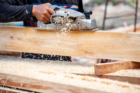 Male carpenter hand using an electric planer on wooden plank in workshop. Woodworker planing timber at construction site with sawdust spreading around. Work tool for carpentry. Lumber industry conceptの写真素材