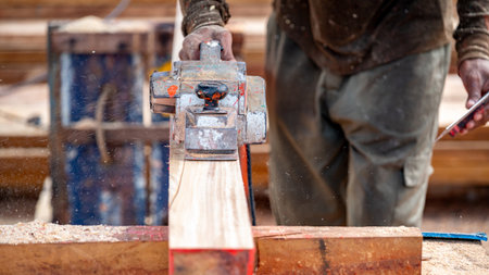 Male carpenter hand using an electric planer on wooden plank in workshop. Woodworker planing timber at construction site with sawdust spreading around. Work tool for carpentry. Lumber industry conceptの写真素材