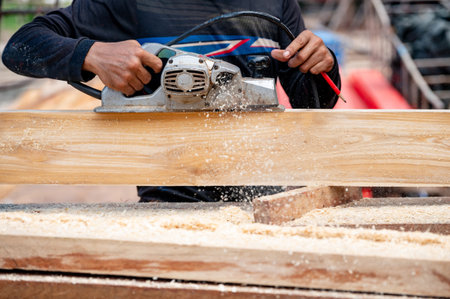 Male carpenter hand using an electric planer on wooden plank in workshop. Woodworker planing timber at construction site with sawdust spreading around. Work tool for carpentry. Lumber industry conceptの写真素材