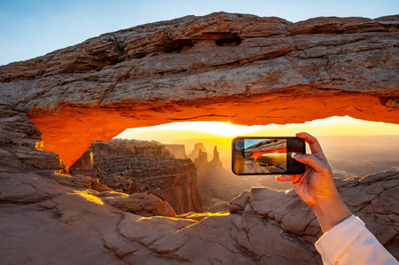 Tourist hand holding smart phone capturing photo of beautiful sunrise at Mesa Arch, a spectacular natural stone arch perched at the edge of a cliff in Canyonlands National Park, Utah, United States.の写真素材