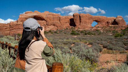 Asian female tourist holding camera taking photo of North Window, arch sandstone rock formation in Arches National Park near Moab in Utah, Southwest USA.の写真素材