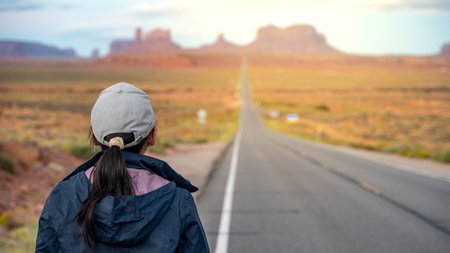 Rear view of Asian female tourist standing off the highway US route 163 looking at the long road view leading to Monument Valley on the Arizona-Utah border, United States. America road trip travelの写真素材