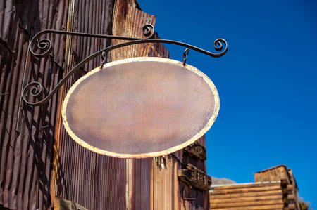 Blank metal antique shop sign. Oval shape vintage signboard with empty space mockup for text design hanging on wrought iron in front of a retro shop with rusty zinc sheet facade in old town.の写真素材