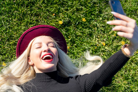 Portrait of a girl with blond hair taking a selfie lying on the grass in a park. Emotion is joy....