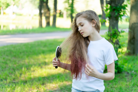 Little girl brushing her hair standing on the grass in the park against the trees. Front view