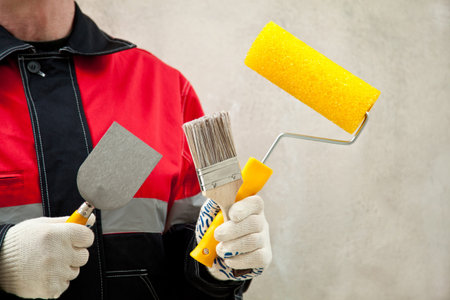 Construction worker in uniform at workplace holding paint roller and brushes; painter with tools