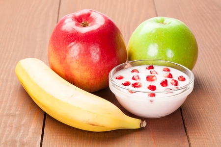 Bowl with natural yoghurt and pomegranate kernels, ripe red and green apples and banana on wooden table; concept of healthy breakfastの写真素材