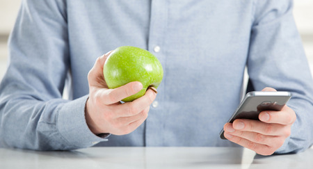 Unrecognizable young man eating fresh green apple and using his smartphoneの写真素材