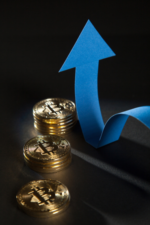 Shiny bitcoins and blue arrow showing up indicating positive trend, black backgroundの写真素材