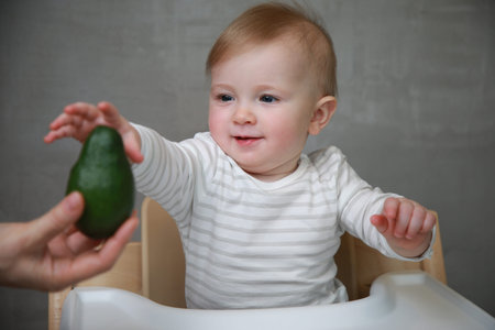 Cute baby boy sitting in highchair and holding avocado in handsの写真素材