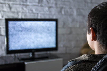 Little boy watching lcd TV in a dark room with loft style brick walls, backside view. Glitched...