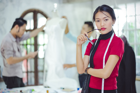 Designer pretty girls in casual clothes and hold the glasses are standing with arms, the cross, look to the camera. His colleagues are working on the rear in the studio.の写真素材