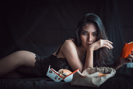Beautiful Sexy woman in sexy lingerie posing on black bedroom and eating a burger looking at camera.  Concept weight loss.の写真素材