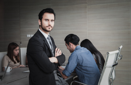 A confident elegant handsome young man standing in front and with his staff in background at officeの写真素材