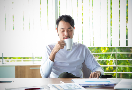 Young man in smart casual wear working drinking some hot drink while sitting at his working place in modern office.の写真素材